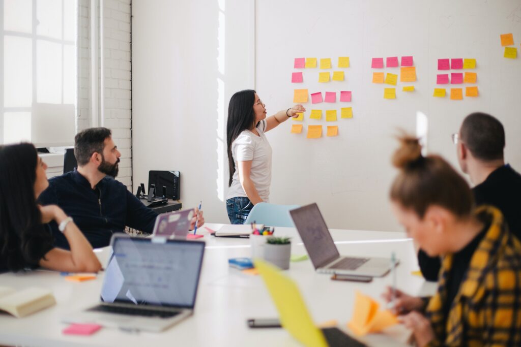 A team in a bright office space engaged in a strategy session with one member presenting ideas on a wall covered in colorful sticky notes, while colleagues with laptops and notebooks sit at a table in discussion.