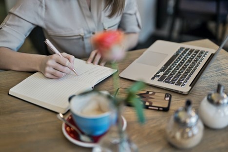 Person writing in a notebook beside a laptop and coffee on a desk.